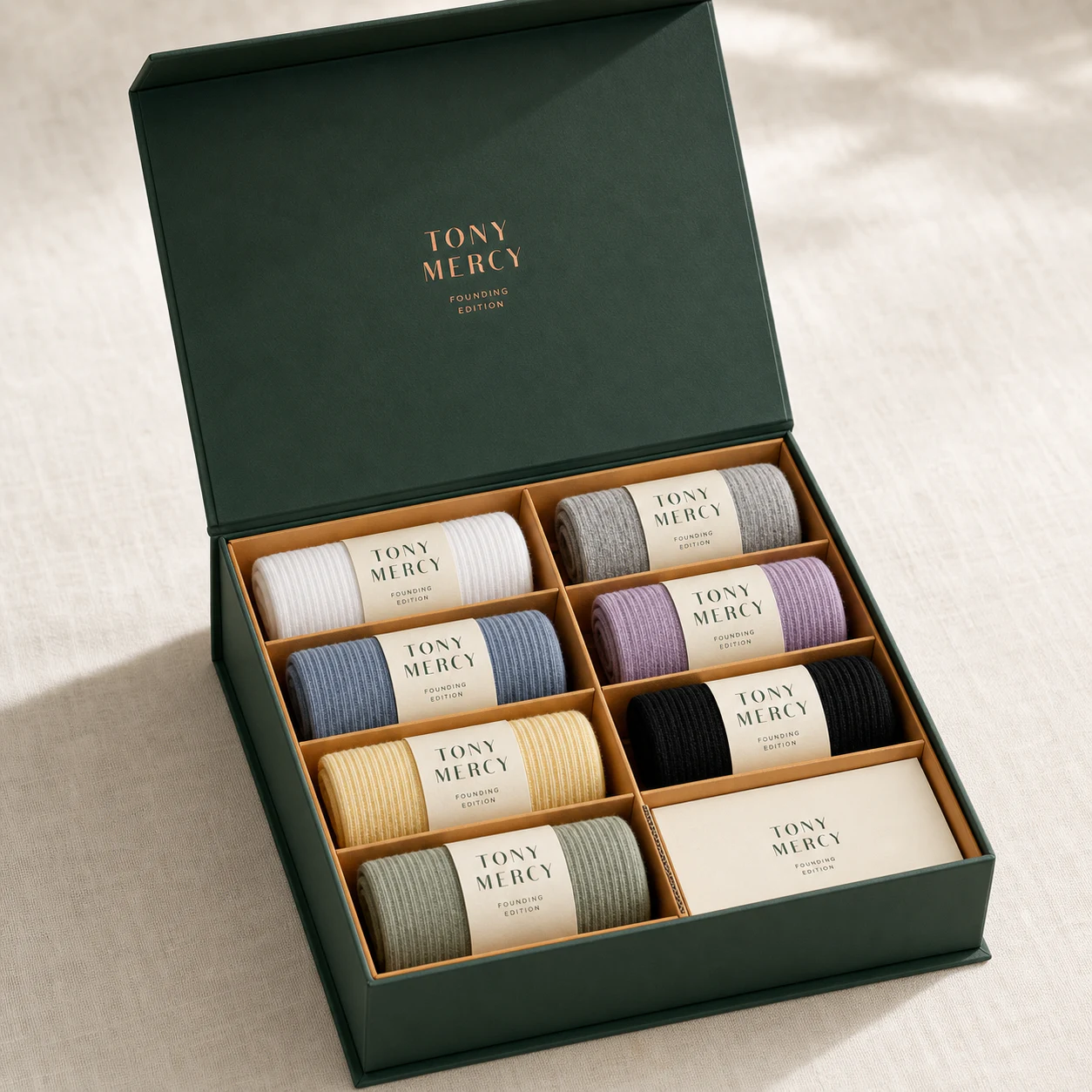 Concept render: open deep-pine magnetic Founding Edition box with seven rolled pairs in paperboard compartments on ivory linen
